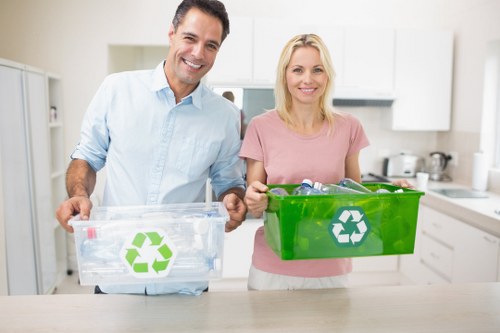 Two individuals, a man and a woman, stand in a bright, modern kitchen and each holds a plastic recycling bin; the man’s clear bin with a green recycling symbol contains various transparent plastic bottles, while the woman’s green bin also with a recycling symbol is filled with other recyclable plastics and packaging. The kitchen features white cabinets, a stainless steel kettle in the background, and a clean, minimalist aesthetic with natural lighting. The scene exemplifies household waste separation and recycling efforts, aligned with rubbish removal services provided by companies such as House Clearance Grays, serving regions around Grays in Essex. The individuals appear engaged in environmentally conscious activity, emphasizing the importance of proper waste segregation for effective rubbish management, especially in residential areas. The detailed view highlights the texture of the plastic bins, the transparency of the bottles, and the bright daylight illuminating the scene, conveying a professional and neutral depiction of domestic recycling practices that support effective waste disposal and clearance solutions in the local community.