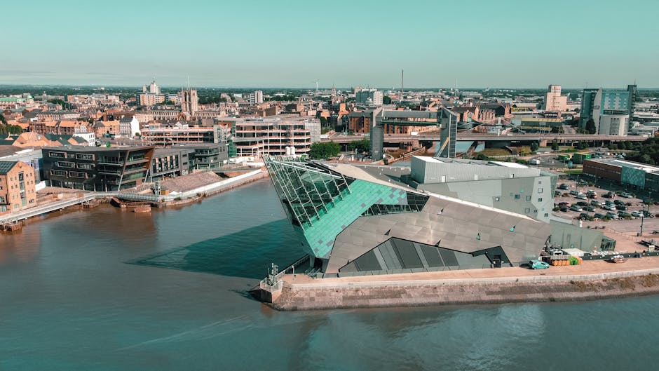 An aerial view of a modern building situated on the edge of a river, featuring a distinctive angular design with a prominent glass facade that reflects light and water. The structure has a combination