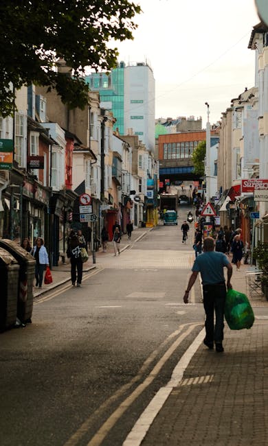 A city street scene in Grays with a mix of traditional and modern buildings lining each side of the road. In the foreground, a man wearing a blue shirt and black trousers is walking away from the came